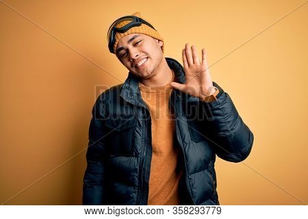 Young brazilian skier man wearing snow sportswear and ski goggles over yellow background Waiving saying hello happy and smiling, friendly welcome gesture