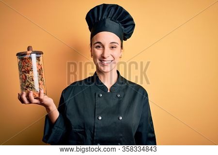 Young beautiful chef woman wearing cooker uniform and hat holding bottle with italian pasta with a happy face standing and smiling with a confident smile showing teeth