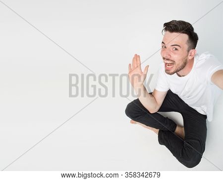 Young Man Isolated Over Background. Guy Sit On Floor And Wave With Hand. Take Selfie Or Picture Alon