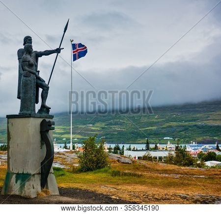 Landnemar Status In Akureyri Iceland Pointing To Icelandic Flag On Cloudy Day. Mountains And Fog In 