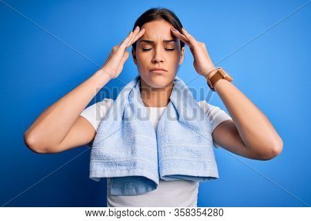Young beautiful brunette sportswoman wearing towel doing exercise over blue background suffering from headache desperate and stressed because pain and migraine. Hands on head.