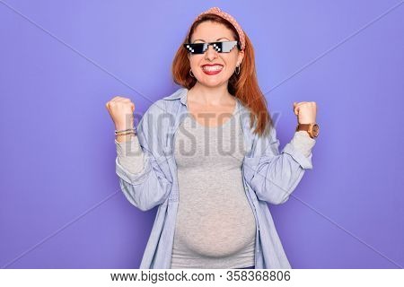 Young redhead pregnant woman expecting baby wearing funny thug life sunglasses very happy and excited doing winner gesture with arms raised, smiling and screaming for success. Celebration concept.