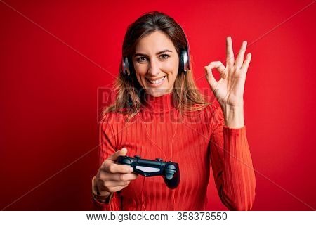 Young beautiful brunette gamer woman playing video game using joystic and headphones doing ok sign with fingers, excellent symbol