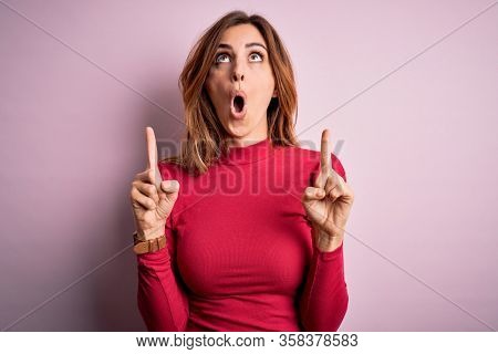 Young beautiful brunette woman wearing casual sweater stnding over pink background amazed and surprised looking up and pointing with fingers and raised arms.