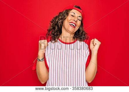 Middle age senior woman wearing baseball equiment over red isolated background very happy and excited doing winner gesture with arms raised, smiling and screaming for success. Celebration concept.