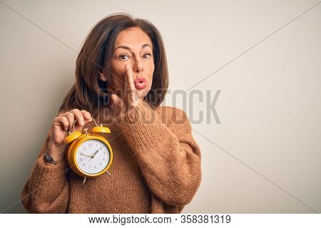 Middle age brunette woman holding clasic alarm clock over isolated background hand on mouth telling secret rumor, whispering malicious talk conversation