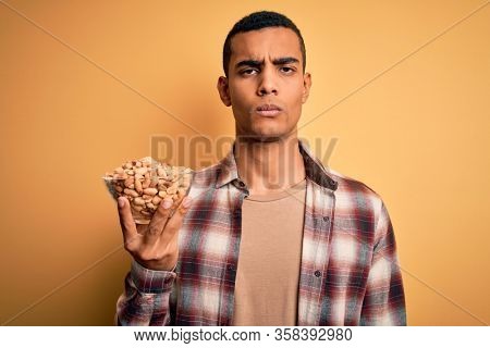 Handsome african american man holding bowl with heathy peanuts over yellow background with a confident expression on smart face thinking serious