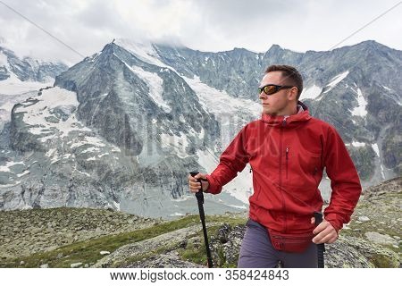 Portrait Of A Man Wearing A Red Jacket And A Backpack, Having A Walk With Hiking Poles In The Mounta