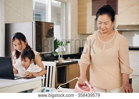 Young Asian Woman Working On Computer With Daughter On Laps When Her Senior Mother Ironing Clothes
