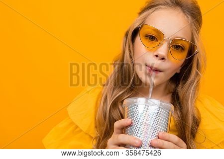Portrait Of Young Caucaian Girl With Long Fair Hair In Yellow Glasses Drinks A Juice And Smiles