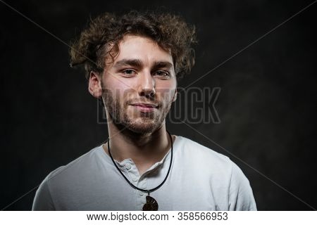 Curly And Hansome Caucasian Man Standing In A Dark Studio On A Grey Background, Wearing Casual White