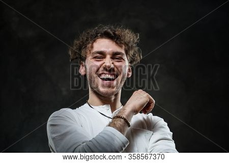 Curly And Hansome Caucasian Man Standing In A Dark Studio On A Grey Background, Wearing Casual White
