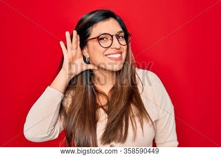 Young hispanic smart woman wearing glasses standing over red isolated background smiling with hand over ear listening an hearing to rumor or gossip. Deafness concept.