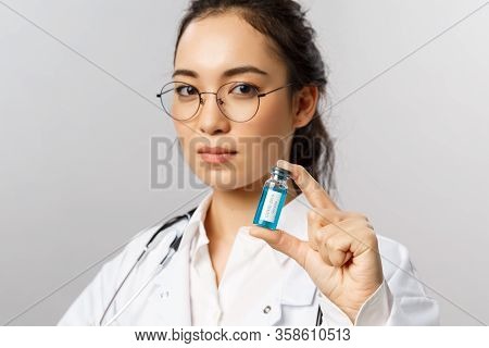 Close-up Portrait Of Serious-looking Young Asian Infectionist, Female Doctor Showing Ampule With Cor