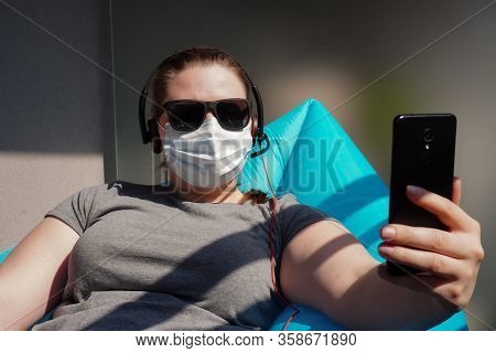 European Girl Blogger In Dark Glasses, Medical Mask, Headphones, Phone Works Remotely Or Conducts Li