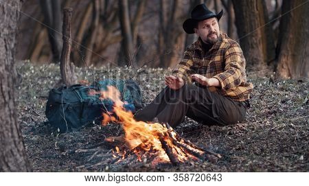 Male Outdoorsman Sitting At Campfire And Warming Hands In Spring Forest Looking Aside