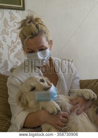 A Woman Hugs Her Cute Dog At Home While Sitting On A Sofa, Wearing A Protective Mask. Concept Of Sta