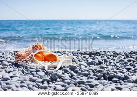 White Cotton Eco String Bag With Oranges On A Rocky Beach. Sea View With A Blue Sky. Zero And Eco Wa