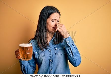Young woman with blue eyes drinking jar of beer standing over isolated yellow background smelling something stinky and disgusting, intolerable smell, holding breath with fingers on nose. Bad smell