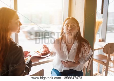 Female Model Touch Stomach And Feels Pain. Young Woman Ate A Lot Of Pizza In A Cafe With Her Friend.
