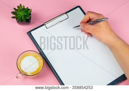 Tablet With White Flattery Next To Which Stands A Green Plant, Hand Holds A Pen And Writes On A Shee