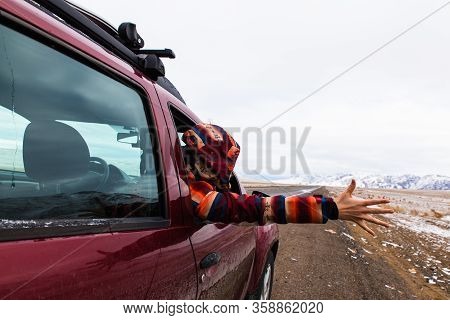 Backview Of Girl In Bright Burgundy Colored Jacket Holding Hand Out Of The Car At Empty Road With Mo