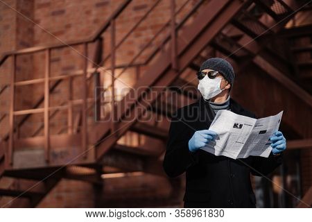 Horiontal Shot Of Pensive Man Poses With Newspaper, Wears Protective Mask And Rubber Gloves, Looks A