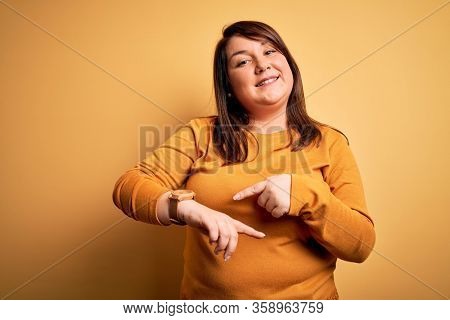 Beautiful brunette plus size woman wearing casual sweater over isolated yellow background In hurry pointing to watch time, impatience, upset and angry for deadline delay