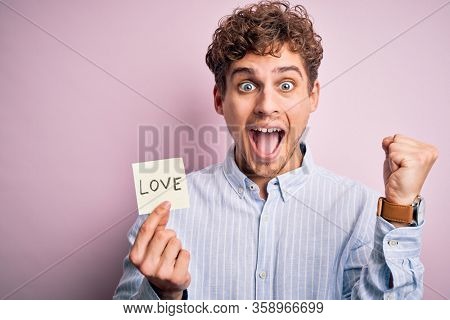 Young blond romantic man with curly hair holdiing paper with love message for valentines day screaming proud and celebrating victory and success very excited, cheering emotion