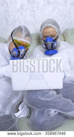 People In Protective Suits And Respirators At Home Using A Laptop. Vertical Shot,