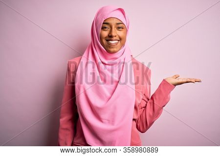 Young African American afro woman wearing muslim hijab over isolated pink background smiling cheerful presenting and pointing with palm of hand looking at the camera.