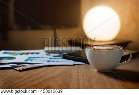 Close Up View, Latte Coffee In White Cup On Wooden Table With Paperwork Of Chart And Tablet Computer
