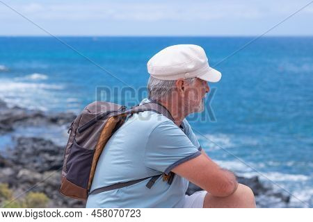Smiling Caucasian Bearded Senior Man With Hat Sitting At The Beach Looking At The Horizon While Enjo