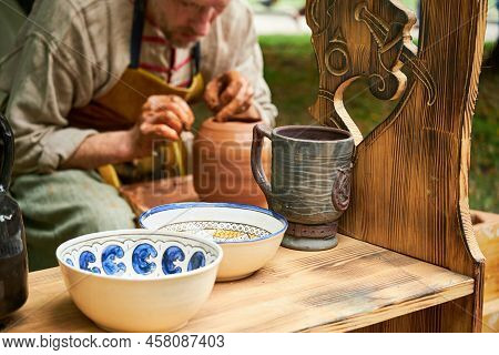 A Man In Medieval Clothes Sits Behind A Vintage Potter Wheel And Makes Dishes Out Of Clay. Times And
