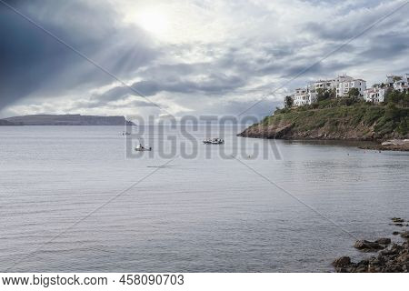Sunset In A Bay With Some White Houses On A Cliff And The Sun Filtering Through The Clouds, Some Boa