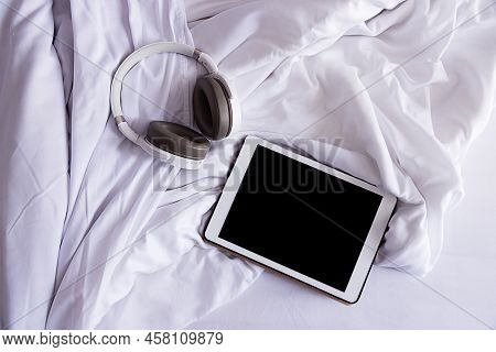A Tablet And Headphones Lie On A Crumpled White Bed, Top View, Close-up