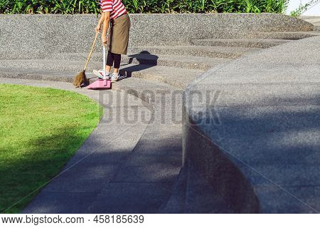 Female Worker Sweeping Yard With Broom Tool And Dustpan In Ornamental Backyard Garden. Woman Wear Ap