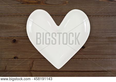 Top View Of A White Heart Shaped Plate Resting On A Modern Rusic Brown Wood Background.