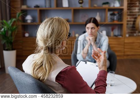 Over Shoulder View Of Female Psychologist Sitting In Armchair, Talking With Upset Woman Patient. Psy