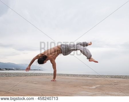 Shirtless Caucasian Man Doing Backflip On Pebble Beach.