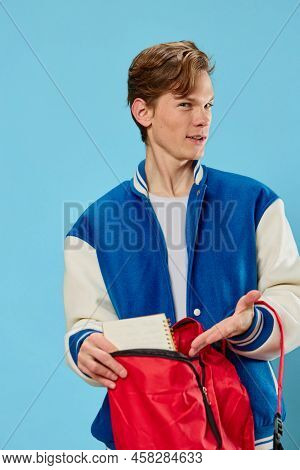 A Pensive Student In A Blue Stylish Bomber Jacket Stands With A Notebook In His Hands And Looks Away