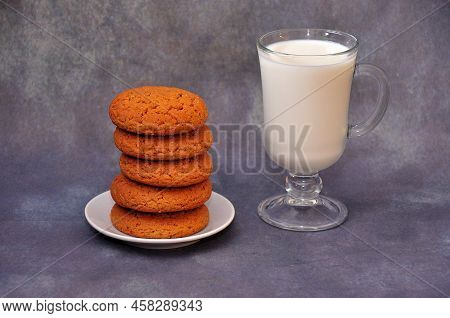 A Stack Of Fresh Oatmeal Cookies On A Saucer And A Glass Of Hot Milk On A Gray Background.