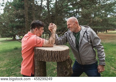 Happy Senior Father With His Adult Son With Down Syndrome Arm Wrestling In Park.