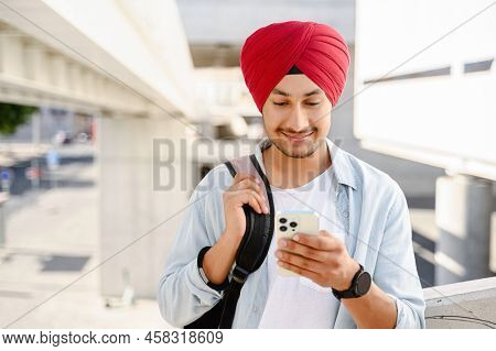 Calm Young Indian Guy In Casual Shirt And Traditional Turban Using Smartphone Outdoors. Male Student
