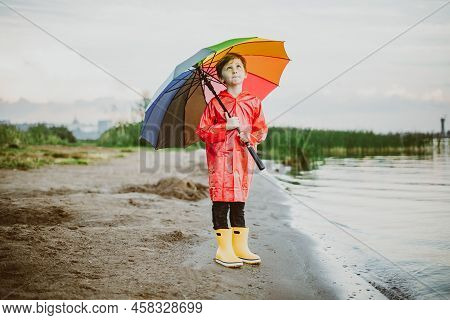 Boy In A Red Raincoat And Yellow Rubber Boots Stands At River Bank And Holding Rainbow Umbrella. Sch