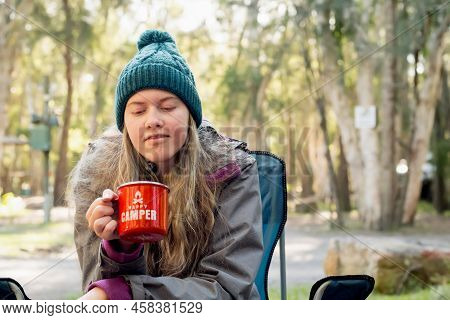 Camping Lifestyle Concept. Teenage Girl Wearing Beanie Hat Sitting In A Camping Chair Near Campfire