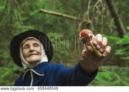 An Elderly Woman Holds A Red Fly Agaric In Her Hands Against The Background Of The Forest. The Old L