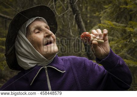 An Elderly Woman Holds A Red Fly Agaric In Her Hands Against The Background Of The Forest. The Old L