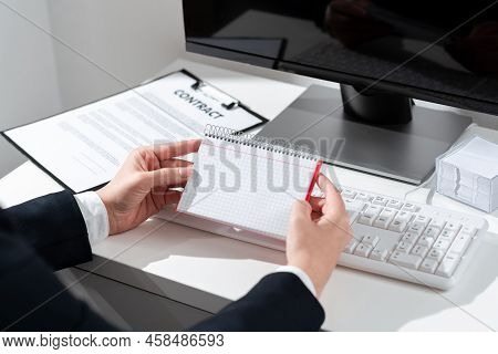 Businesswoman Holding Important Message Wtitten On Notebook On Desk With Computer, Clipboard And Not