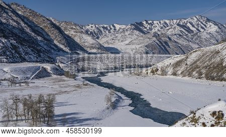 Turquoise Ice-free River Winds Among The Icy Shores. Bare Trees In The Valley. Picturesque Mountains